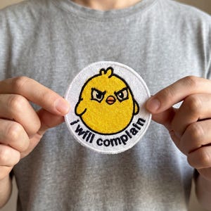 May include: A round, white patch with a yellow chick illustration and the text "I will complain" in black. The chick has an angry expression. The patch is held up by a person.