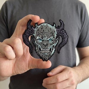 May include: Embroidered patch featuring a detailed demon skull design with large horns. The patch has a gray and black color scheme with teal accents in the eyes. The patch is held up in front of a gray t-shirt.