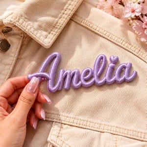 Könnte beinhalten: Ein hellvioletter Stickaufnäher mit dem Namen Amelia in Schreibschrift, ergänzt durch ein kleines Herz über dem Buchstaben i. Der Aufnäher ist auf einer beigen Cordjacke mit sichtbaren Knöpfen und Nähten angebracht.
