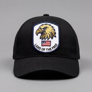 May include: Black baseball cap featuring an embroidered patch with a golden eagle, the American flag, and the text "LAND OF THE FREE". The cap has a curved brim and a structured crown, suitable for casual wear.