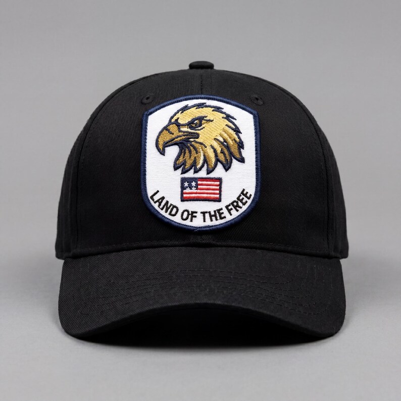 May include: Black baseball cap featuring an embroidered patch with a golden eagle, the American flag, and the text "LAND OF THE FREE". The cap has a curved brim and a structured crown, suitable for casual wear.