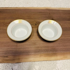 May include: Two white and gold rimmed bowls with a gold emblem are displayed on a wooden surface. The bowls have a simple, elegant design, with a gold band around the rim and a gold emblem on the side.