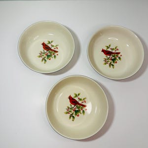 May include: Three off-white ceramic bowls with a red cardinal bird and holly berry design. Each bowl has a black rim and a glossy finish. The bowls are arranged in a triangular pattern on a white surface.