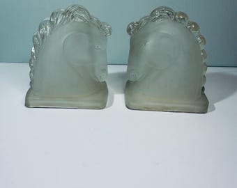 May include: Two frosted glass bookends, each shaped like a horse head. The horse heads are facing opposite directions, with detailed manes and facial features. The bookends are a light, translucent color.