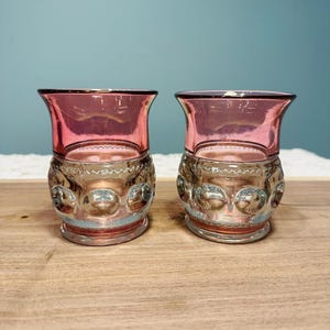 May include: Two vintage pink and clear glass tumblers. The tumblers have a decorative, textured base with a series of raised circles. The rims are a darker pink, and the glasses sit on a wooden surface.