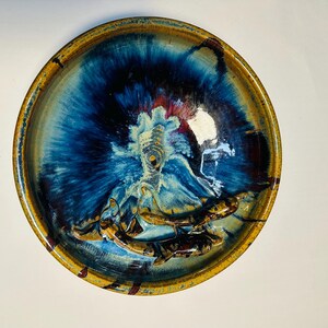 May include: A round ceramic bowl with a vibrant, glazed finish. The bowl features a deep blue center that radiates outwards, transitioning to lighter blues and a golden-brown rim. The interior has a raised, textured design.