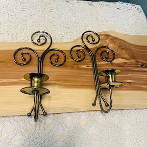 May include: A pair of wall-mounted candle holders crafted from polished brass-toned metal. Each piece features a vertical spine with three decorative scrolls at the top and a circular platform designed to support a single pillar candle.