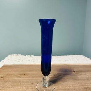 May include: A tall, cobalt blue glass vase with a flared top and a clear, round base. The vase features etched floral designs and a clear, bulbous stem. The vase is approximately 10 inches tall.