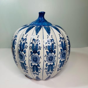 May include: A white and blue ceramic pumpkin-shaped decorative jar. The jar features a blue floral pattern and a blue stem. The jar is a decorative item for the home.