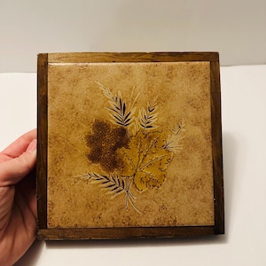 May include: Square wooden coaster with a light brown, textured surface. The coaster features a botanical design of leaves and sprigs in shades of brown and tan. The coaster is framed by a dark brown wooden border.