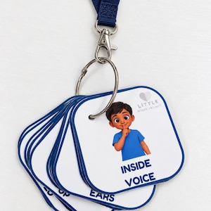 Può includere: Un set di carte quadrate bianche con bordi blu fissate a un anello di metallo e un cordino blu scuro. La carta superiore presenta l'illustrazione di un bambino con capelli scuri e maglietta blu che porta un dito alle labbra con il testo.