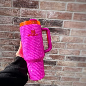 May include: A pink Stanley tumbler with a handle, covered in sparkling rhinestones. The lid is orange, and the handle is a darker pink. The Stanley logo is visible on the side of the tumbler. The tumbler is held against a brick wall.