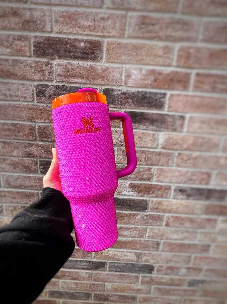 May include: A pink Stanley tumbler with a handle, covered in sparkling rhinestones. The lid is orange, and the handle is a darker pink. The Stanley logo is visible on the side of the tumbler. The tumbler is held against a brick wall.