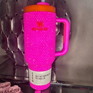 May include: A bright pink Stanley Quencher H2.0 tumbler with a bedazzled exterior. The tumbler has an orange lid and handle, a white straw, and a label that reads "40oz | 1.18L All Day Hydration".