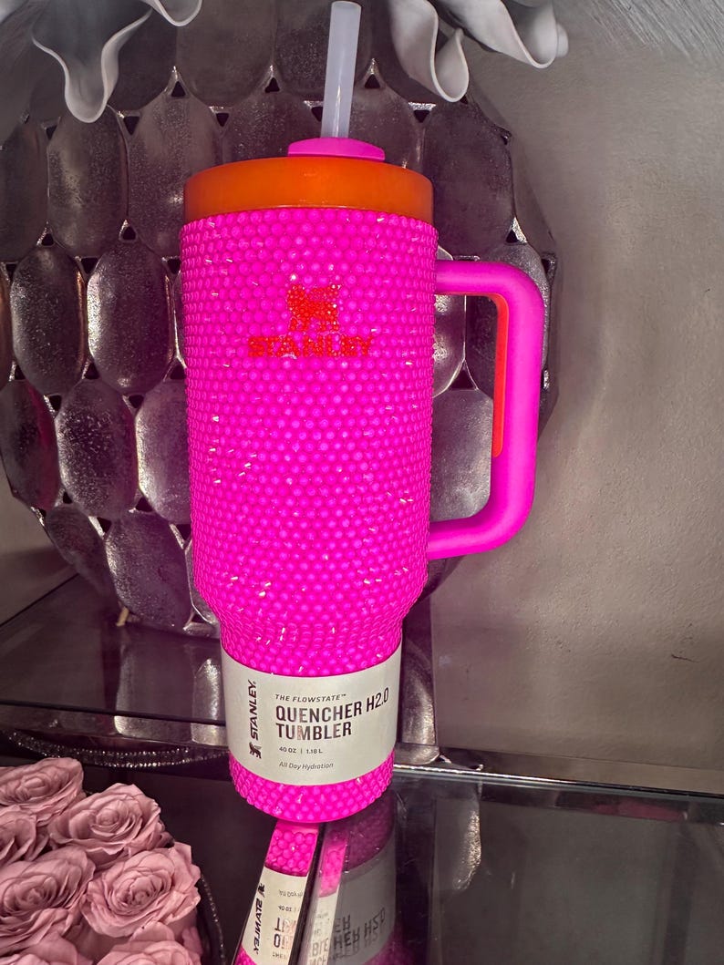 May include: A bright pink Stanley Quencher H2.0 tumbler with a bedazzled exterior. The tumbler has an orange lid and handle, a white straw, and a label that reads "40oz | 1.18L All Day Hydration".