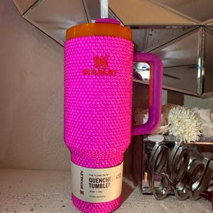 May include: A hot pink Stanley Quencher 40 oz tumbler covered in rhinestones. The tumbler has an orange lid, a purple handle, and a white straw. The text "THE FLOWSTATE QUENCHER 420 TUMBLER" is printed on a white label.