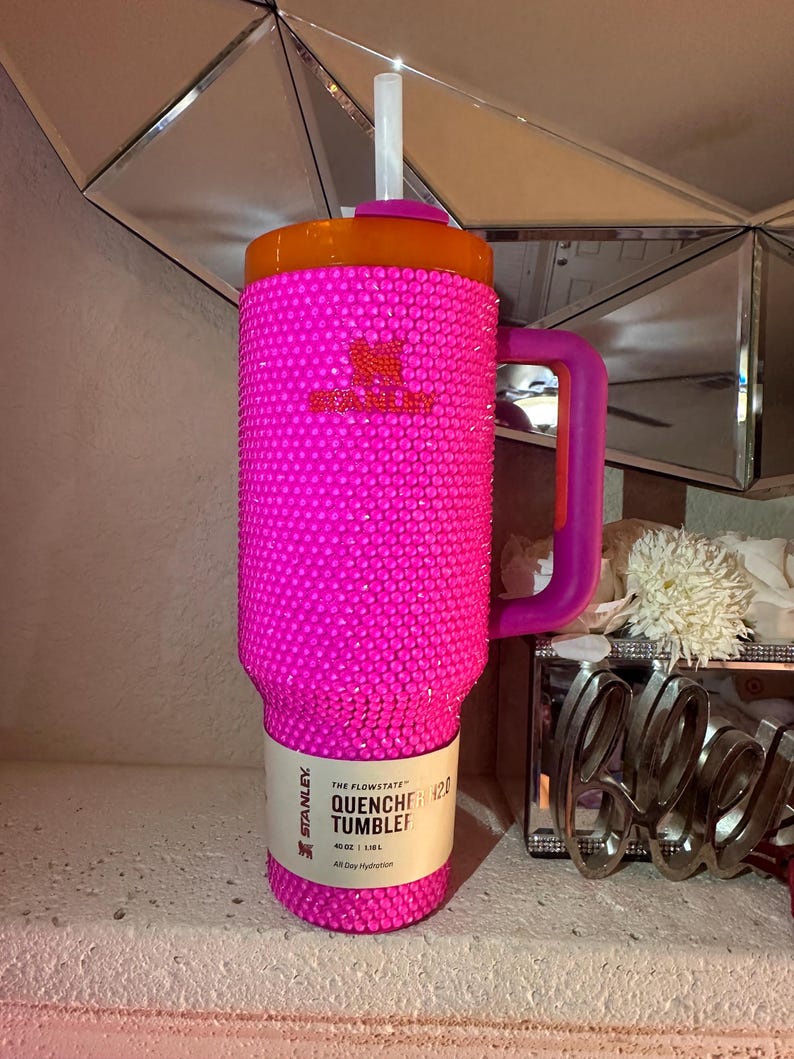 May include: A hot pink Stanley Quencher 40 oz tumbler covered in rhinestones. The tumbler has an orange lid, a purple handle, and a white straw. The text "THE FLOWSTATE QUENCHER 420 TUMBLER" is printed on a white label.