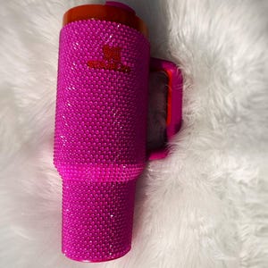 May include: A vibrant pink tumbler covered in sparkling rhinestones. The tumbler has a matching pink handle and a red lid. A small red logo is visible on the side. This insulated drinkware is perfect for hot or cold beverages.