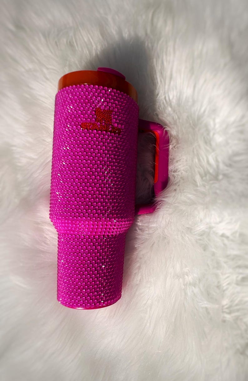 May include: A vibrant pink tumbler covered in sparkling rhinestones. The tumbler has a matching pink handle and a red lid. A small red logo is visible on the side. This insulated drinkware is perfect for hot or cold beverages.