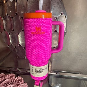 May include: A hot pink Stanley Quencher H2.0 Tumbler with a handle and a straw. The tumbler is covered in small, sparkling embellishments. The lid is orange. The tumbler holds 40 oz, or 1.18 L.