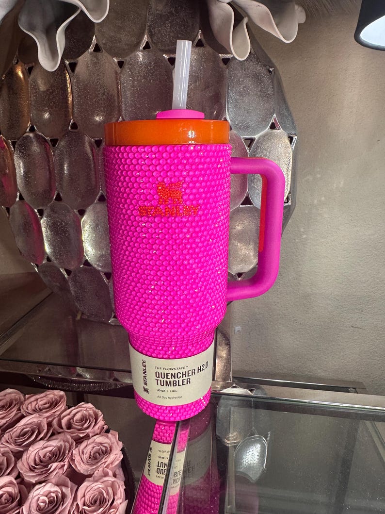 May include: A hot pink Stanley Quencher H2.0 Tumbler with a handle and a straw. The tumbler is covered in small, sparkling embellishments. The lid is orange. The tumbler holds 40 oz, or 1.18 L.