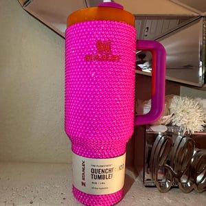 May include: A hot pink Stanley Quencher tumbler with a bedazzled exterior. The tumbler has a purple handle and an orange lid with a white straw. The label reads "THE FLOWSTATE QUENCHER TUMBLER".