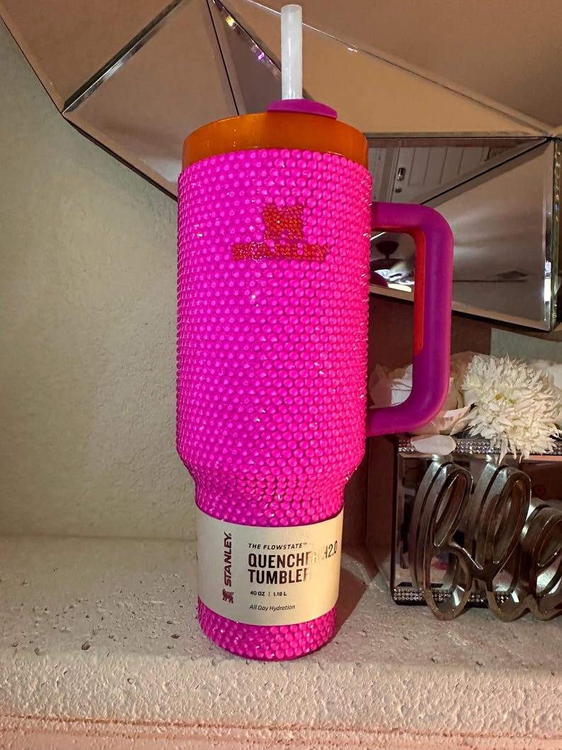 May include: A hot pink Stanley Quencher tumbler with a bedazzled exterior. The tumbler has a purple handle and an orange lid with a white straw. The label reads "THE FLOWSTATE QUENCHER TUMBLER".
