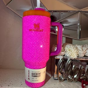 May include: A vibrant pink Stanley Quencher 40 oz tumbler, covered in sparkling embellishments. The tumbler has an orange lid and a purple handle. The label reads "QUENCHER 2.0 TUMBLER". A white straw extends from the lid.