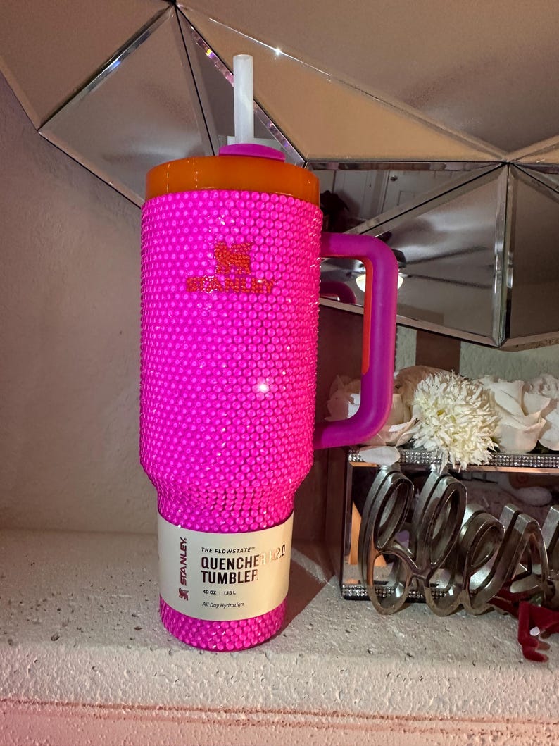 May include: A vibrant pink Stanley Quencher 40 oz tumbler, covered in sparkling embellishments. The tumbler has an orange lid and a purple handle. The label reads "QUENCHER 2.0 TUMBLER". A white straw extends from the lid.