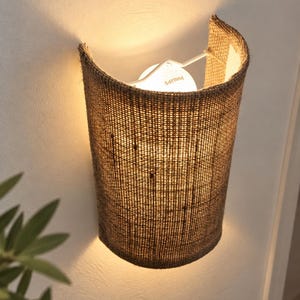 Op de afbeelding: Een halfcilindervormige wandlamp met een kap van gestructureerd bruin jute. Het armatuur bevat een witte Philips-lamp die een warm licht uitstraalt en zowel naar boven als naar beneden op de muur achter de lamp schijnt.