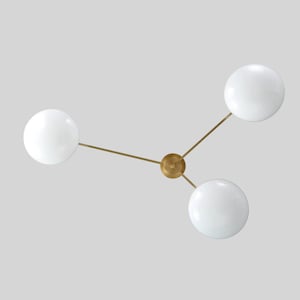 Op de afbeelding: Een moderne plafondlamp met drie witte bolvormige kappen. De kappen zijn verbonden met een centrale messingkleurige naaf door dunne, rechte armen. De lamp staat tegen een lichtgrijze achtergrond.