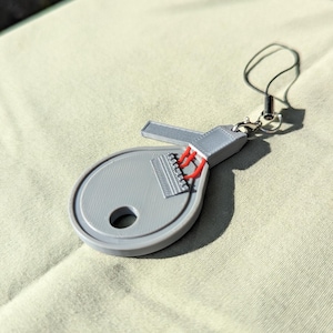 Peut inclure: Un porte-clés gris imprimé en 3D en forme de luth, avec des détails de cordes rouges. Le porte-clés a un petit trou et une lanière noire attachée. Le luth repose sur une surface vert clair.