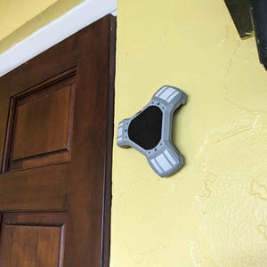 May include: A gray and black wall-mounted object with a unique, geometric design. The object is attached to a yellow wall, near a dark brown wooden door. The object has a central black section and gray arms with white accents.