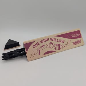 Może przedstawiać: Gadżet One Wish Willow w beżowym trójkątnym pudełku z fioletowym tekstem i grafiką. Pudełko zawiera napisy "ONE WISH WILLOW", "AMAZE YOUR FRIENDS!" oraz "You only get ONE WISH". Z pudełka wystaje czarny, teksturowany przedmiot przypominający gałąź.