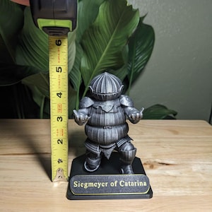 May include: A silver-colored figurine of Siegmeyer of Catarina, approximately 5 inches tall, stands on a black base. The figurine is of a knight in armor. The base has a gold-colored nameplate with the text "Siegmeyer of Catarina."