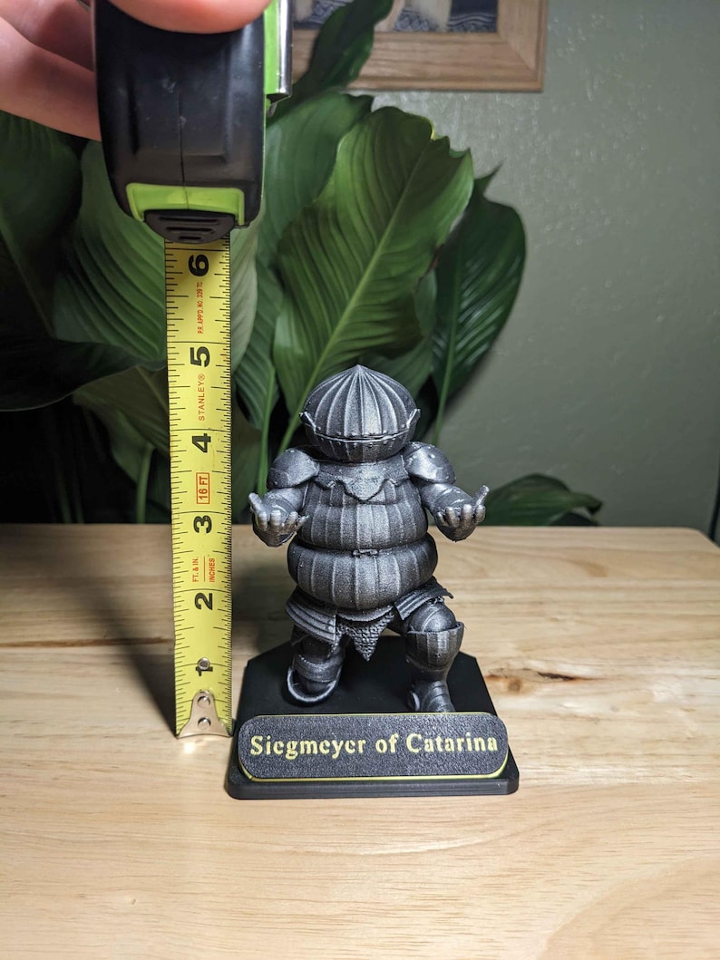 May include: A silver-colored figurine of Siegmeyer of Catarina, approximately 5 inches tall, stands on a black base. The figurine is of a knight in armor. The base has a gold-colored nameplate with the text "Siegmeyer of Catarina."