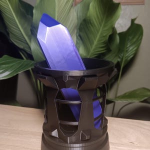 May include: A faceted, purple crystal stands in a black, geometric holder. The holder has a cylindrical base and an open, cage-like design. The crystal is a deep violet color and appears to be made of a translucent material. The background features large green leaves.