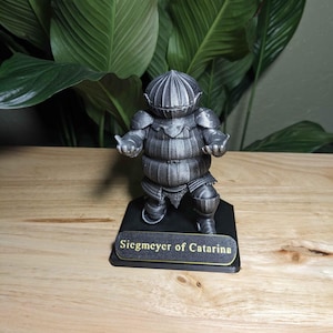 May include: A detailed, gray figurine of a knight in full armor, standing on a black base. The base has the text "Siegmeyer of Catarina" in gold lettering. The figurine is set against a backdrop of large green leaves.