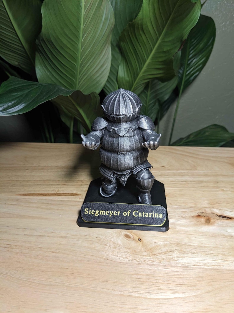 May include: A detailed, gray figurine of a knight in full armor, standing on a black base. The base has the text "Siegmeyer of Catarina" in gold lettering. The figurine is set against a backdrop of large green leaves.