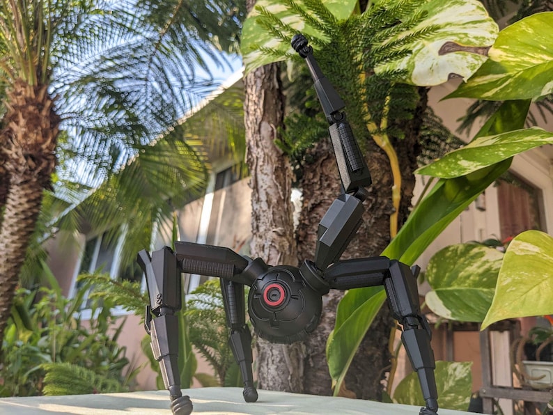 Pode incluir: Um brinquedo rob&oacute;tico preto, mec&acirc;nico, semelhante a uma aranha, com pernas articuladas e um corpo esf&eacute;rico central. O rob&ocirc; tem um detalhe vermelho e preto semelhante a um olho e est&aacute; situado em um cen&aacute;rio de folhagem verde exuberante e palmeiras.