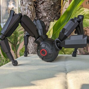Pode incluir: Um brinquedo de aranha mec&acirc;nica preta com pernas articuladas e um corpo central. O corpo apresenta um detalhe circular vermelho. O brinquedo est&aacute; posicionado sobre uma superf&iacute;cie de cor clara, com vegeta&ccedil;&atilde;o ao fundo.