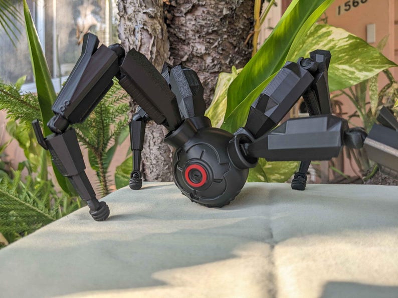 Pode incluir: Um brinquedo de aranha mec&acirc;nica preta com pernas articuladas e um corpo central. O corpo apresenta um detalhe circular vermelho. O brinquedo est&aacute; posicionado sobre uma superf&iacute;cie de cor clara, com vegeta&ccedil;&atilde;o ao fundo.