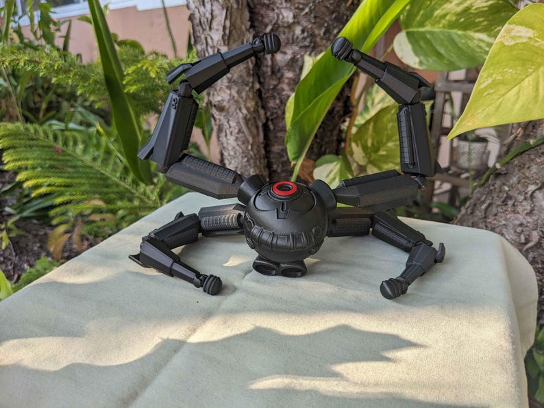 Pode incluir: Um brinquedo de aranha mec&acirc;nica preta articulada com um corpo central e m&uacute;ltiplas pernas articuladas. O corpo central tem um detalhe circular vermelho. O brinquedo est&aacute; posicionado em uma superf&iacute;cie verde clara, com um fundo natural de plantas e um tronco de &aacute;rvore.