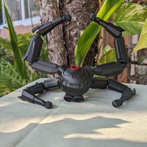 Pode incluir: Um modelo mec&acirc;nico preto, semelhante a uma aranha, com pernas e bra&ccedil;os articulados. O corpo central &eacute; esf&eacute;rico com um detalhe circular vermelho. O modelo &eacute; exibido em uma superf&iacute;cie de cor clara, com um fundo natural de folhagem verde e um tronco de &aacute;rvore.