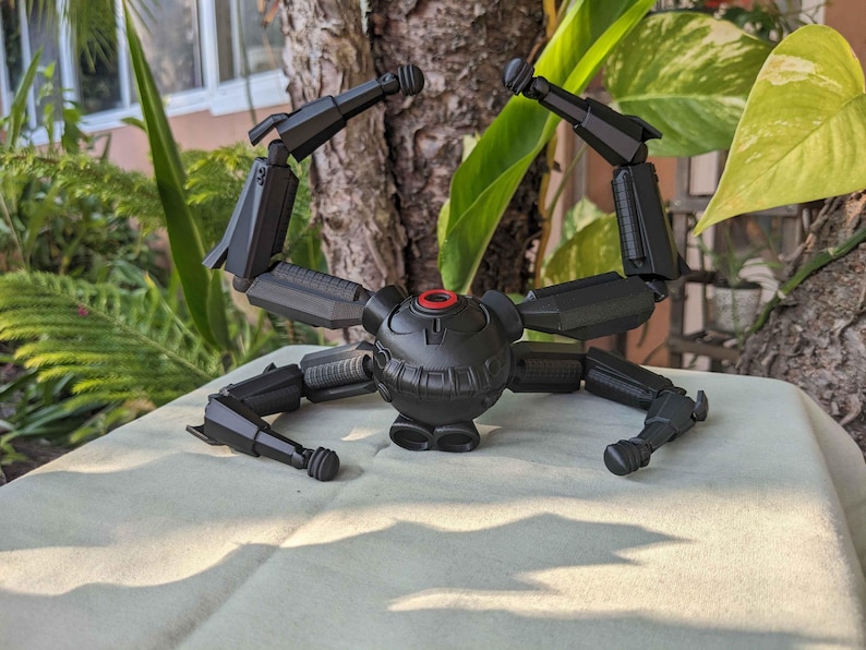 Pode incluir: Um modelo mec&acirc;nico preto, semelhante a uma aranha, com pernas e bra&ccedil;os articulados. O corpo central &eacute; esf&eacute;rico com um detalhe circular vermelho. O modelo &eacute; exibido em uma superf&iacute;cie de cor clara, com um fundo natural de folhagem verde e um tronco de &aacute;rvore.