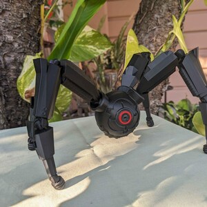 Pode incluir: Um modelo mec&acirc;nico preto semelhante a uma aranha com pernas articuladas e um corpo central com um detalhe circular vermelho. O modelo est&aacute; posicionado sobre uma superf&iacute;cie de cor clara, com um fundo natural de folhagem e casca de &aacute;rvore.
