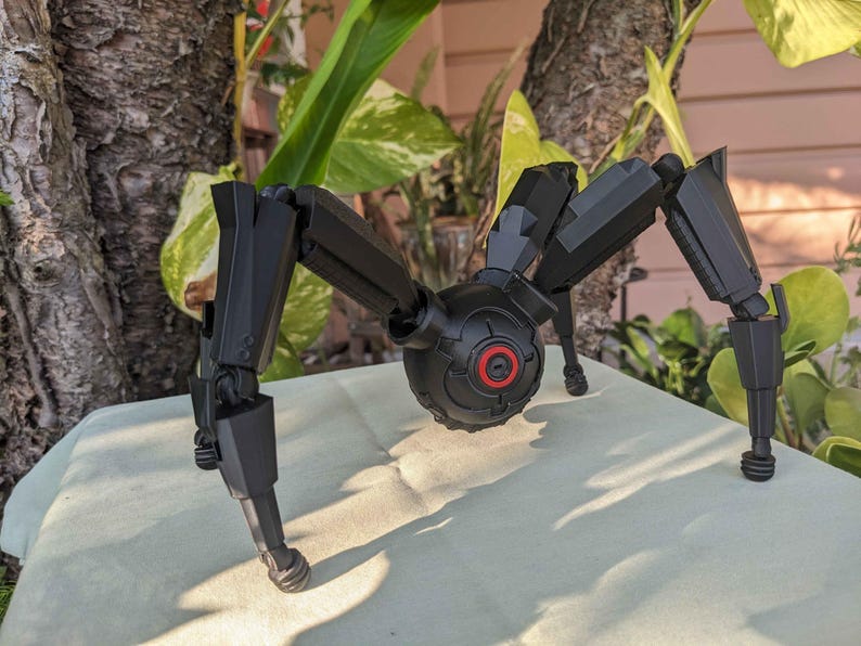 Pode incluir: Um modelo mec&acirc;nico preto semelhante a uma aranha com pernas articuladas e um corpo central com um detalhe circular vermelho. O modelo est&aacute; posicionado sobre uma superf&iacute;cie de cor clara, com um fundo natural de folhagem e casca de &aacute;rvore.