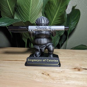 May include: A silver-colored figurine of a knight holding a black and white Sharpie marker. The figurine is on a black base with the text "Siegmeyer of Catarina" in gold lettering. The background features large green leaves.