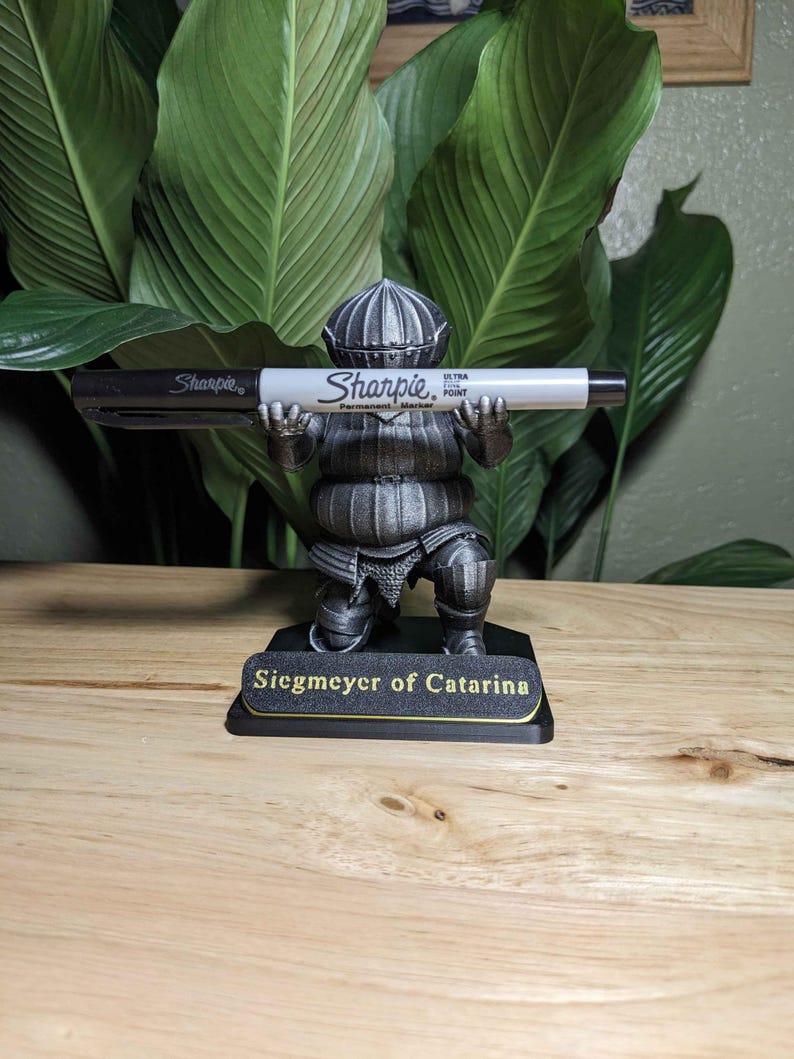May include: A silver-colored figurine of a knight holding a black and white Sharpie marker. The figurine is on a black base with the text "Siegmeyer of Catarina" in gold lettering. The background features large green leaves.