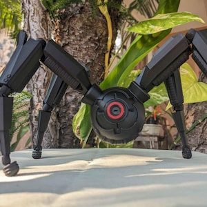 Pode incluir: Um modelo preto, mec&acirc;nico, semelhante a uma aranha, com pernas articuladas e um corpo central. O corpo apresenta um design circular com um detalhe vermelho. O modelo est&aacute; sobre um fundo natural com folhagem.